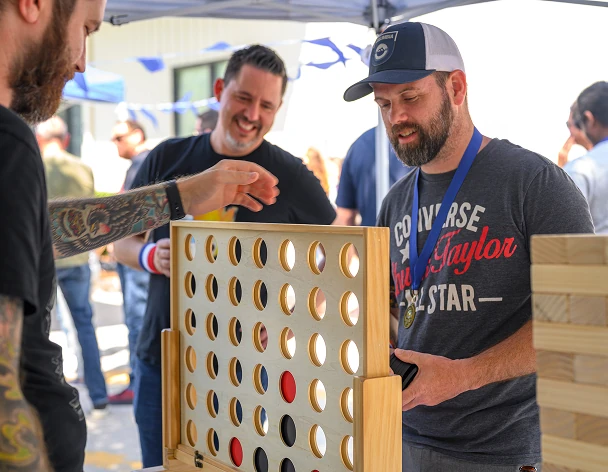 Three people playing a large outdoor Connect Four game with red and black checkers, with one person dropping a piece, set under a tent with blue decorations.