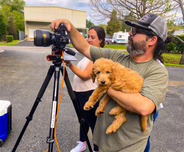 A man with a beard and cap holding a fluffy golden puppy, standing next to a woman adjusting a camera on a tripod in an outdoor setting with a building and trees in the background.