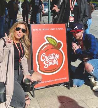 Two people posing in front of a red sign with a peach logo reading 'Creative South' and the slogan 'Come as friends, leave as family,' with a crowd and urban background.